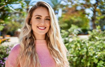A woman with long blonde hair wearing a pink sweater stands outdoors, smiling at the camera with greenery and sunlight in the background.